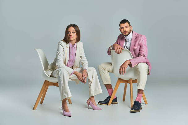 trendy business couple in pastel formal wear sitting in armchairs on grey, minimalistic fashion - Photo, Image