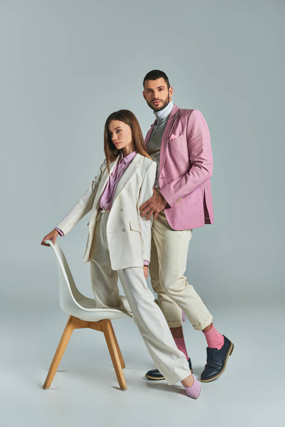 junges elegantes Paar in pastellfarbener formaler Kleidung posiert mit Sessel auf grauem, modernem Businessstil - Foto, Bild