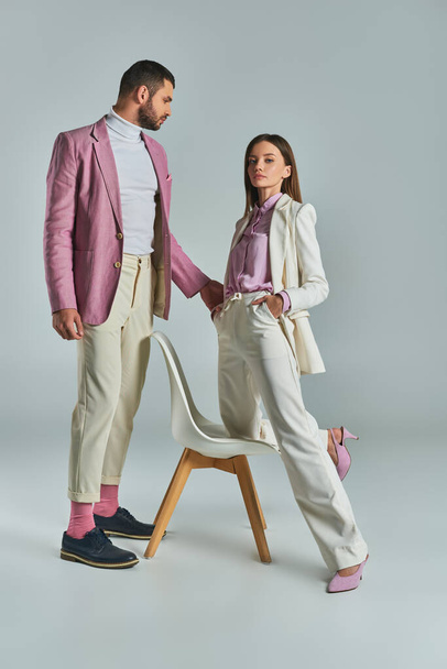 femme confiante en costume blanc avec les mains dans les poches près de l'homme en blazer lilas et fauteuil sur gris - Photo, image