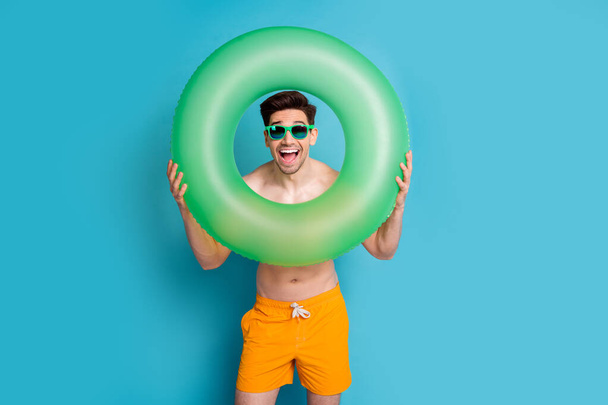 Photo portrait of handsome young guy inflatable ring frame dressed stylish yellow shorts isolated on blue color background summer vacation. - Foto, afbeelding