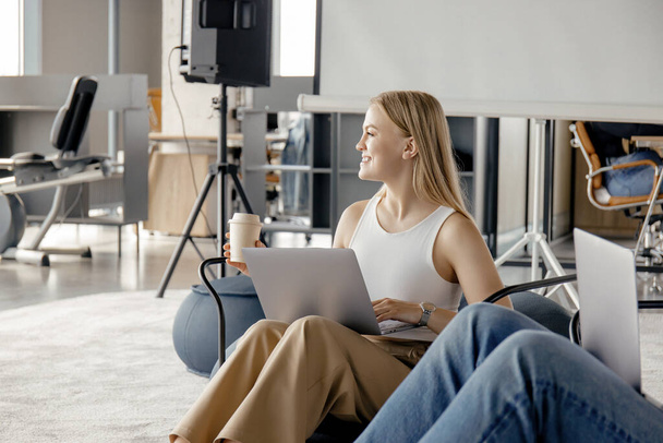 Uma jovem feliz está trabalhando em um laptop com um café na mão enquanto se senta em uma cadeira confortável - Foto, Imagem