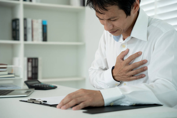 рука тримає груди з симптомами серцевого нападу, азіатський чоловік важко працює має біль у грудях, викликаний серцевими захворюваннями, витоком, розширенням коронарного серця, натисканням на груди з болючим виразом - Фото, зображення