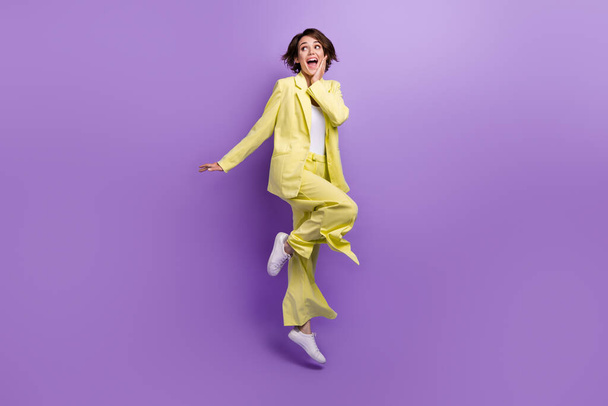 Full length photo of shiny excited woman dressed yellow jacket jumping high looking empty space isolated purple color background. - Photo, image