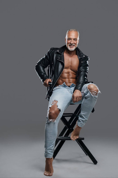Masculine and stylish male model with striking physique, grey beard, and bare chest, wearing black leather jacket and ripped jeans, sitting on a studio chair against a neutral grey backdrop - Photo, Image