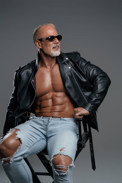 Pohledný mužský model se stylovým šedým vousem se slunečními brýlemi, v černé kožené bundě a roztrhaných džínách, odhalující jeho svalnatou postavu a holý trup při pózování na židli ve studiu - Fotografie, Obrázek