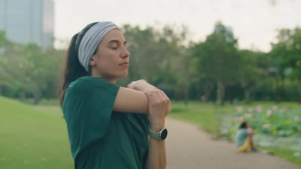 Spor kıyafetli genç bir kadının parkta antrenmandan önce kollarını, bileklerini ve omuzlarını esneterek ısınırken orta kavisli pozu. - Video, Çekim