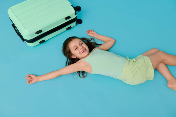 Caucasian mischievous little child girl in stylish wear, with bright turquoise suitcase, lying on blue background, expressing positive emotions, looking at camera. Travel and summer holidays concept. - Photo, Image