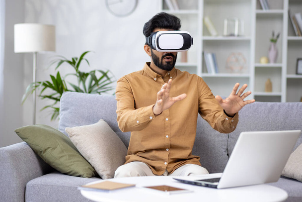 Zaskoczony poważny człowiek w okularach wirtualnej rzeczywistości przeglądając kurs w wirtualnym spotkaniu siedząc na kanapie w salonie w domu. - Zdjęcie, obraz