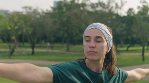 Střední zblízka záběr ženy ve sportovním oblečení a čelenka cvičení válečník pózovat s pažemi nataženými při cvičení jógy v parku - Záběry, video