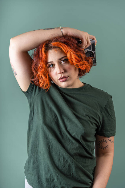 Modern ve dövmeli, ikili olmayan kızıl saçlı, eski bir fotoğraf makinesi ve askeri renkleri olan bir kız.. - Fotoğraf, Görsel