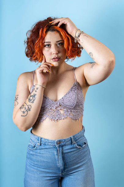 Moderni tatuoitu ei-binary punapää tyttö katselee sinua sinisellä taustalla - Valokuva, kuva