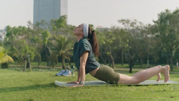 Jonge sportieve vrouw doet vinyasa yoga en dan rusten in kinderen poseren op trainingsmat in het park tijdens de ochtend outdoor praktijk - Video