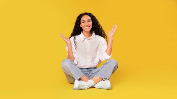 Jovem árabe feliz em Casual com cabelos longos sentado no chão e Gesturing com as mãos, sorrindo para a câmera, expressando emoções positivas e publicidade Oferta agradável em fundo amarelo. Panorama - Foto, Imagem