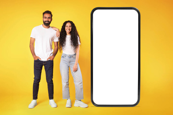 Casual middle eastern couple standing near large smartphone with blank screen, looking at camera, advertising offer or mobile app for phone, mockup, over yellow background. Full length shot - Photo, Image