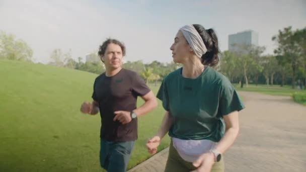 Średni długi slow motion shot młodej pary w odzieży sportowej biegającej w parku i rozmawiającej podczas treningu na świeżym powietrzu rano - Materiał filmowy, wideo