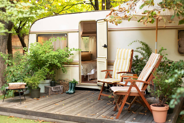 Cozy Campsite on caravan or camper van in forest. Trailer of mobile home stands in garden in camping. Backyard with RV house with garden furniture. Two deckchairs near outside caravan trailer.  - Foto, Imagen