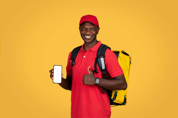 alegre millennial afro-americano cara mensageiro com mochila mostrar telefone com tela vazia, polegar para cima, isolado no fundo do estúdio laranja. Site de aprovação, recomendação de aplicativo para entrega em domicílio - Foto, Imagem