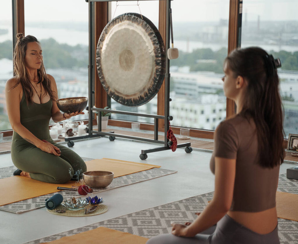 Serene moment captured as girls engage in spiritual practices and yoga, accompanied by the soothing sound of Tibetan singing bowl, against backdrop of picturesque cityscape and breathtaking sunset - Photo, Image