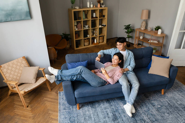 Asyalı genç çift dijital tablet kullanıyor, online film izliyor, evdeki koltukta birlikte dinleniyorlar, yüksek açılı. Eşler hafta sonunun tadını çıkarıyor, İnternet 'te sörf yapıyor. Gadget Kullanımı, İnternet Teknolojisi - Fotoğraf, Görsel