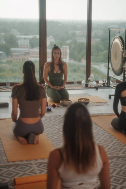Spor giyimli kızlar şehir manzarasına bakan bir dairede grup seansı sırasında yoga ve meditasyon yapıyorlar. - Fotoğraf, Görsel