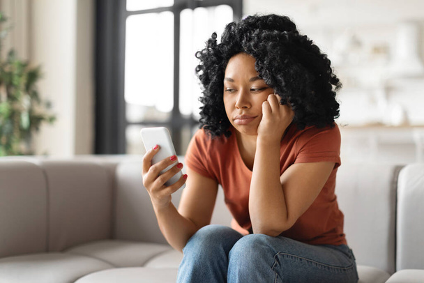 Αναστατωμένη νεαρή Αφροαμερικανή γυναίκα κάθεται στον καναπέ και κοιτάζοντας την οθόνη Smartphone, βαριέται Millennial μαύρη γυναίκα περιμένει σημαντική κλήση ή sms ενώ χαλαρώνει στο σπίτι, δωρεάν χώρος - Φωτογραφία, εικόνα