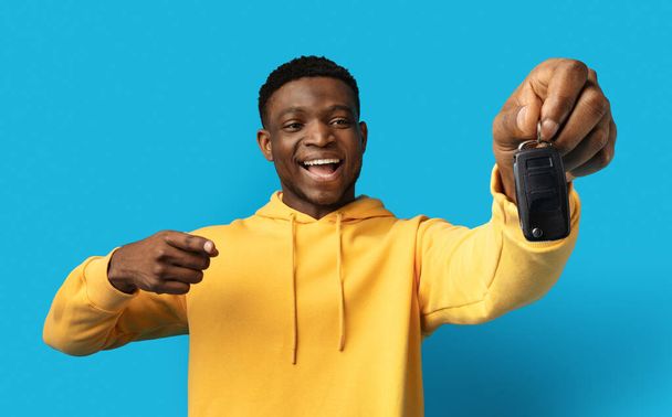 Happy excited vivid driver young black man pointing at key from new car in his hand, isolated on blue background, studio shot, copy space. Driving, car leasing buying concept - Fotografie, Obrázek