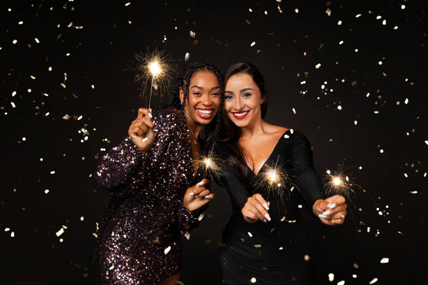 Gelukkige multi-etnische vriendinnen twee jonge vrouwen dragen mooie elegante outfits houden bengal lichten, lachen onder confetti schittert, vieren Nieuwjaar 2024 samen, geïsoleerd op zwarte achtergrond - Foto, afbeelding