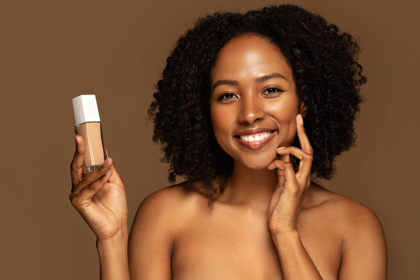 Lächelnd sexy attraktive halbnackte junge schwarze Frau mit glühend perfekter Haut, die Flasche mit Schönheitsprodukt Foundation oder Concealer zeigt, isoliert auf braunem Studiohintergrund, Nahaufnahme - Foto, Bild