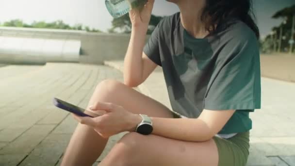 Tilt up πλάνο της νεαρής γυναίκας σε αθλητικά ρούχα και κεφαλόδεσμο κάθεται σε εξωτερικούς χώρους στην πόλη, πόσιμο νερό από το μπουκάλι και τη χρήση smartphone, ενώ ξεκουράζεται μετά το πρωινό τρέξιμο - Πλάνα, βίντεο