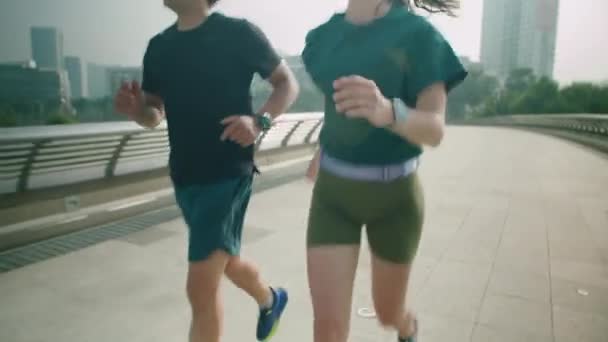 午前中に一緒に運動しながら,街の歩行者橋に沿って一緒に走っている2人のカップルをトリミング - 映像、動画