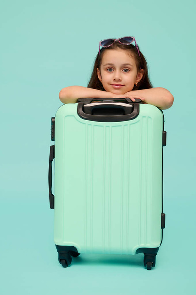 Красивая кавказская маленькая девочка, путешественник, турист, пассажир, собирается на выходные отдыха, уверенно глядя в камеру, позируя с голубым чемоданом, изолированные над студийным фоном. Копирование рекламного пространства - Фото, изображение