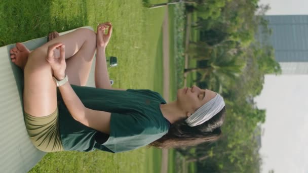 Tiro vertical de mujer joven deportiva sentada en pose de loto y manos en mudras mientras practica meditación de yoga por la mañana en el parque - Imágenes, Vídeo