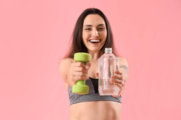 ピンクの背景に水とダンベルのボトルが付いているスポーティな若い女性 - 写真・画像