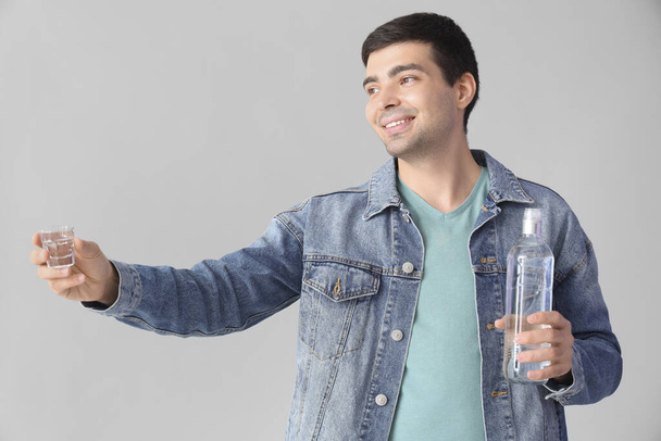 Νεαρός άνδρας με ποτήρι και μπουκάλι βότκα σε ελαφρύ φόντο - Φωτογραφία, εικόνα