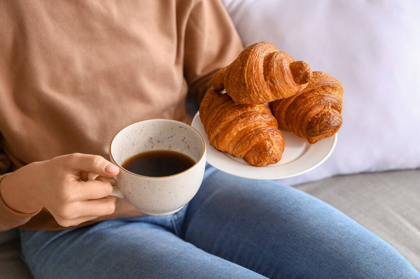 Young woman with tasty croissants and cup of coffee sitting on sofa in living room - Photo, Image