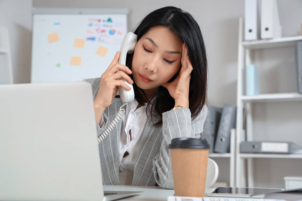 Κουρασμένη Ασιάτισσα επιχειρηματίας που μιλάει τηλεφωνικώς και εργάζεται στο γραφείο - Φωτογραφία, εικόνα