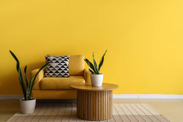 Άνετη πολυθρόνα, τραπεζάκι και φυτά εσωτερικού χώρου κοντά στον κίτρινο τοίχο - Φωτογραφία, εικόνα