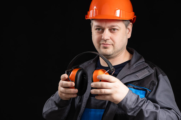 Builder in a protective helmet with noise-cancelling headphones in his hands on a black background, copy space for text. - Photo, Image