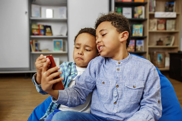 Hermanos viendo entretenido vídeo en el teléfono inteligente cuando se sienta en la silla de la bolsa de frijoles en casa. Tecnología inalámbrica - Foto, imagen