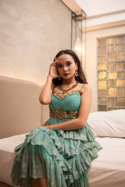 uma mulher asiática com um colar de ouro e brincos está posando lindamente em um quarto de hotel antes do jantar à noite - Foto, Imagem