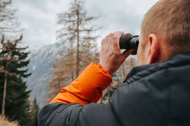 Człowiek z lornetką Oglądanie gór. Wędrówki w górach w sezonie wiosennym.mężczyzna w ciepłej kurtce patrzy przez lornetkę na górski krajobraz. Spacerując w górach - Zdjęcie, obraz
