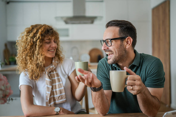 młody para kaukaski mężczyzna i kobieta mąż i żona cieszyć się filiżankę herbaty lub kawy rano w domu szczęśliwy uśmiech codziennie rano rutyna prawdziwych ludzi - Zdjęcie, obraz