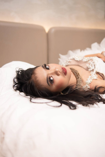 una mujer asiática con el pelo negro mordiéndose los dedos con una expresión cachonda mientras duerme en una cama blanca por la noche - Foto, Imagen