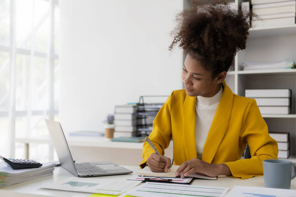 Portret van een gelukkige Afro-Amerikaanse zakenvrouw die werkt met behulp van een laptop computer en papierwerk met een werkplan voor financiële documenten, belastingen en financiële adviseurs. - Foto, afbeelding