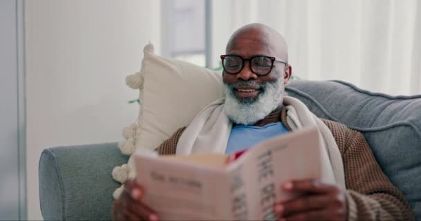Sonrisa, hombre negro maduro y libro de lectura en casa en el sofá de la sala de estar. Feliz, novedoso y africano en el salón para relajarse, estudiar literatura y conocimiento de la historia de fantasía en la jubilación hobby en casa. - Metraje, vídeo
