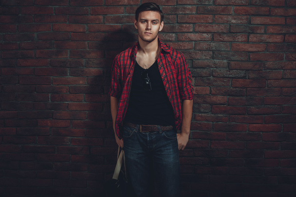 Хипстер серьезный человек случайная одежда расстегнутая рубашка и джинсы в джинсах стоя возле кирпичной стены рука в кармане, глядя на камеру
 - Фото, изображение