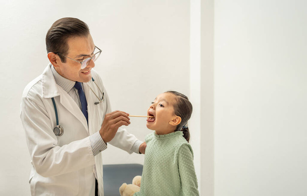 Latijnse dokter die de keel van het meisje controleert met een tongspatel. Spaanstalige lachende kinderarts onderzoekt kind zere keel, kind bij de dokter - Foto, afbeelding