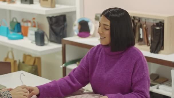 Mujer joven asistente de tienda en el mostrador hablando con la mujer rubia contemporánea comprar suéter en sala de exposición de diseño - Imágenes, Vídeo