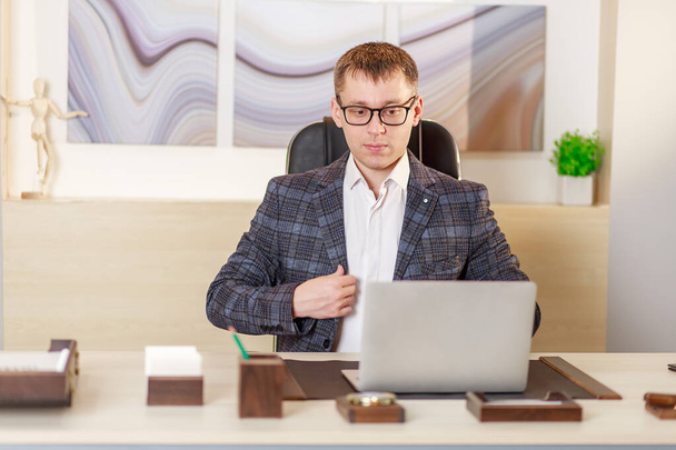 Um homem de negócios senta-se no escritório em uma grande mesa na frente de um laptop e trabalha on-line, bate-papo por vídeo, comunicação sobre momentos de trabalho na Internet, trabalho do escritório, o conceito de trabalho remoto. - Foto, Imagem