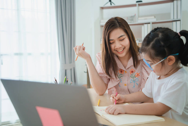 Azjatycka matka uczy dziewczynkę nosić okulary robi w domu za pomocą laptopa i pisać notatki w notatniku do nauki online na drewnianym stole w salonie w domu. Uczenie się przez całe życie. - Zdjęcie, obraz